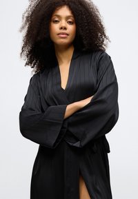 Μαύρη σατέν ρόμπα με λεία υφή, κάθετες ρίγες, φαρδιά μανίκια και βαθύ V-λαιμόκομμα; διαθέτει δέσιμο στη μέση και πλευρική σχισμή.