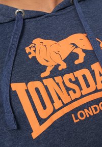 Tengerészeti kapucnis pulóver narancssárga oroszlán grafikával és "Lonsdale London" felirattal. Felszerelt húzózsinóros kapucni, puha, texturált anyaggal.