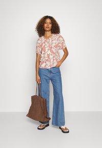 Top branco com padrão colorido em espiral, mangas curtas bufantes e detalhe de amarração no pescoço; combinado com jeans flare azuis e uma bolsa de palha marrom.
