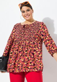 Blusa nera con motivo floreale in rosa e arancione, corpino arricciato, maniche tre quarti, abbinata a pantaloni rossi e una borsa nera.