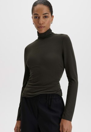 Femme portant un haut à col roulé vert foncé ajusté et un pantalon noir, debout devant un fond clair uni.