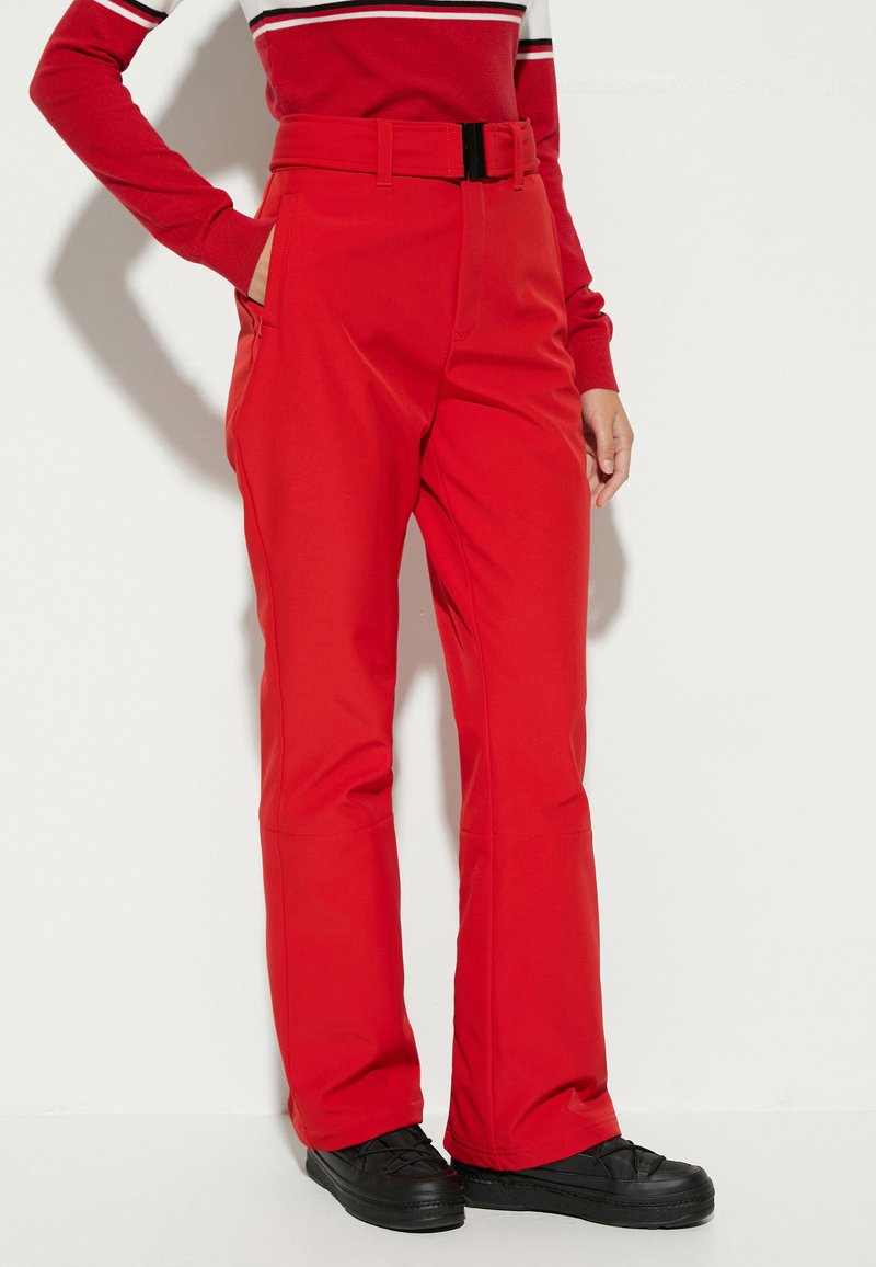 Pantalones de nieve rojos ajustados con un cinturón, bolsillos laterales y una tela ligeramente texturada. Combinados con zapatos negros de cordones.