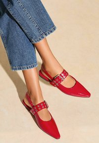 Le ballerine rosse a punta slingback presentano una finitura lucida in vernice, dettagli decorativi con fibbie e occhielli e una suola nera a profilo basso.