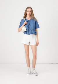 Denim peplum tipo marškiniai su trumpomis rankovėmis ir mazgo detalėmis, derinami su baltais šortais. Modelis laiko mažą baltą rankinę ir dėvi baltus sportbačius.