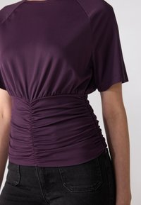 Lilla T-skjorte med korte ermer og samlet midje, laget av mykt stoff med en jevn tekstur. Kombinert med sorte høytlivsjeans.