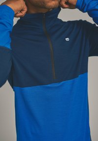Blue long-sleeve top featuring a zip collar. The top has a two-tone design with a dark navy upper and a lighter blue lower section.