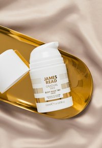 Bottiglia a pompa bianca con dettagli dorati etichettata "James Read Gradual Tan Sleep Mask Tan Face", posizionata su un vassoio ovale dorato contro uno sfondo di tessuto morbido.