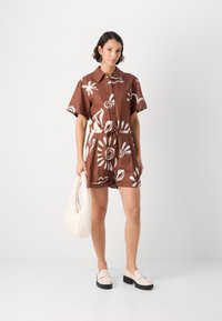 Romper in lino marrone con bottoni, decorato con motivi floreali bianchi, maniche corte e vita regolabile con coulisse. Abbinato a scarpe chiare e una borsa.
