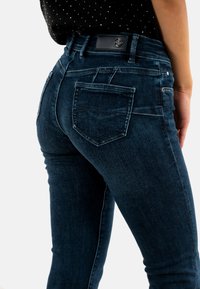 Vaqueros de mezclilla azul oscuro con corte ajustado. Las características incluyen bolsillos traseros y un parche de cuero con marca en la cintura. Textura suave.