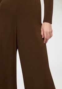 Jumpsuit marrone a gamba larga in tessuto testurizzato. Il primo piano mostra una manica aderente e una mano appoggiata sul materiale liscio.