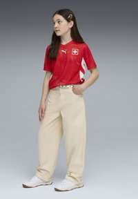 Κόκκινη αθλητική φανέλα με λευκές λεπτομέρειες και έμβλημα της σημαίας της Ελβετίας, συνδυασμένη με φαρδιά μπεζ παντελόνια και λευκά αθλητικά παπούτσια. Μακριά σκούρα μαλλιά με κλιπ.