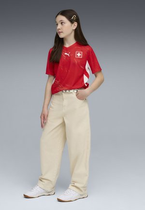 Rood sportief shirt met witte accenten en Zwitserse vlagembleem, gecombineerd met losse beige broek en witte sneakers. Lang donker haar met haarclip.