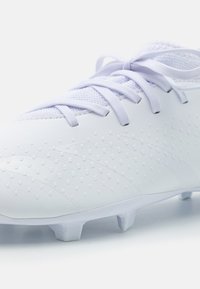 Scarpa da calcio bianca con una parte superiore testurizzata caratterizzata da piccole protuberanze, lacci sottili e un design a profilò basso. Include sei tacchetti modellati sulla suola.