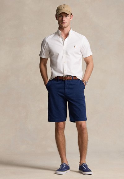 Camisa branca de manga curta com botões, calções azul-marinho, cinto castanho trançado, boné bege e ténis azuis. Design simples e limpo com estilo casual.