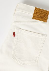 Jean en velours côtelé blanc Levi's avec poche arrière visible, étiquette rouge Levi's et patch de marque en cuir marron sur la ceinture.