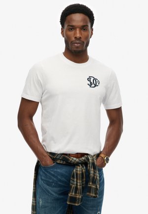 Mężczyzna w białej koszulce z logo, niebieskich dżinsach i koszuli w kratę zawiązanej wokół talii, stoi z rękami w kieszeniach.