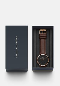 Reloj redondo con esfera negra, manecillas doradas y correa de cuero marrón, presentado en una caja azul marino con letras doradas.