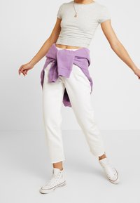 Osoba oblečená v bílých vysokých teniskách, bílých kalhotách, šedém zkráceném topu a fialové mikině uvázané kolem pasu, pózující s roztaženýma rukama.