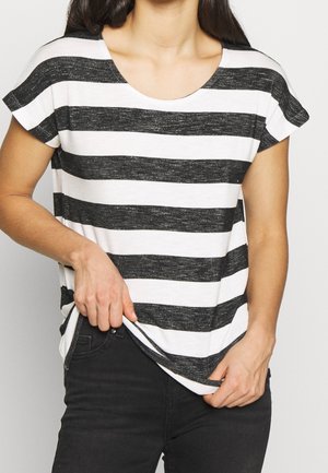 Nainen, joka pukeutunut mustavalkoiseen vaakaraitapaita lyhythihainen, vetää paidan helmaa mustiin housuihin pukeutuneena yksiväristä taustaa vasten.