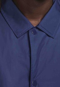 Mörkblå jacka med krage, tryckknappar, slät textur och synlig sömnad på kragen och placket.