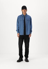 Camicia di denim leggera blu con due tasche frontali, abbinata a una t-shirt nera, jeans neri e stivali neri. Il modello indossa un cappellino nero.
