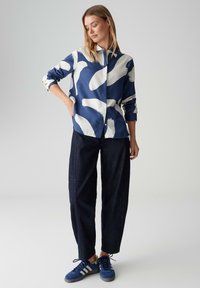 Bluse in Marineblau mit großem weißem Pinselstrichmuster, durchgeknöpft, lange Ärmel. Kombiniert mit locker sitzender dunkler Jeanshose und blauen Turnschuhen.