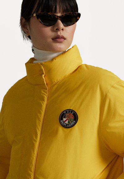 Geel pufferjack met hoge kraag, voorzien van een rond geborduurd logo. Model draagt een schildpad-bril met cat-eye lenzen en een witte coltrui.