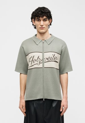 Jeune homme aux cheveux bouclés portant une chemise à manches courtes gris clair zippée avec le texte "Holzweiler" sur la poitrine et un pantalon noir.