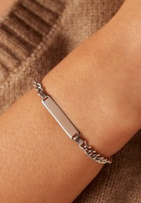 Silver metal bracelet with a flat rectangular centerpiece and a chain link design, resting on a skin-toned wrist. Smooth and shiny texture.