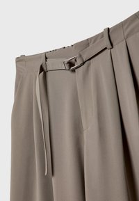 Pantalon taille haute taupe avec passants pour ceinture, fine ceinture en tissu nouée avec une boucle rectangulaire argentée, et plis devant sur fond clair.