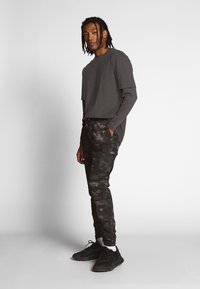 Tumši pelēki garām piedurknēm T-krekls kombinācijā ar melniem rakstainiem kravas biksēm, kurām ir kabatas. Valkā ar melnām sporta apavām un baltām īsām zeķēm.