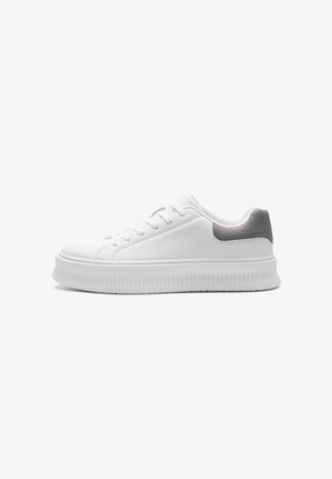 Biały niski sneaker z grubą żeberkowaną podeszwą i szarą teksturowaną łatą na pięcie, widziany z boku na jednolitym tle.