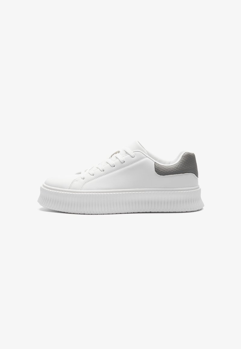 Baskets basses blanches avec une semelle épaisse nervurée et un patch gris texturé au talon, vues de côté sur un fond uni.