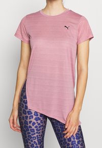 Woman wearing pink asymmetrical Puma athletic top with mesh sleeves and blue leopard-print leggings, standing against plain background.