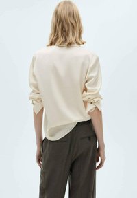 Beige Bluse mit langen Ärmeln und umgeschlagenen Manschetten, aus leichtem Material. Die Rückseite hat einen abgerundeten Saum und einen subtilen Glanz. Kombiniert mit olivgrünen Hosen.