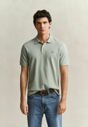 Junger Mann mit kurzen lockigen Haaren, der ein hellgrünes Poloshirt und blaue Jeans trägt, steht vor einem schlichten Hintergrund.