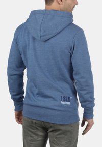Blauwe hoodie met een trekkoordcapuchon, ribgebreide manchetten en zoom, met een achterlogo dat "ISD Sinds 1993" in donkerblauwe borduursel weergeeft.