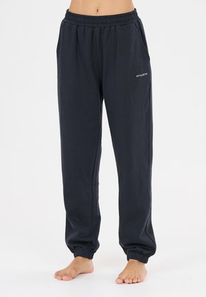 Person iført sorte joggebukser med elastisk talje og ankelmanchetter samt "ATHLECIA" logo på øverste lår, stående barfodet på hvid overflade.