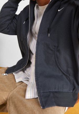 Persoon die een zwarte hoodie met rits draagt over een gestreept shirt en lichtbruine corduroy broek, zittend met één hand dichtbij het hoofd opgeheven.