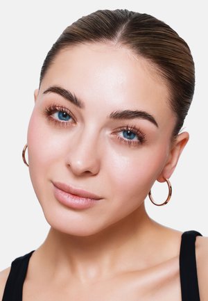 Close-up van een persoon met een gladde huid, blauwe ogen en lichte make-up, die kleine gouden ringoorbellen draagt en een strakke haarbun heeft.
