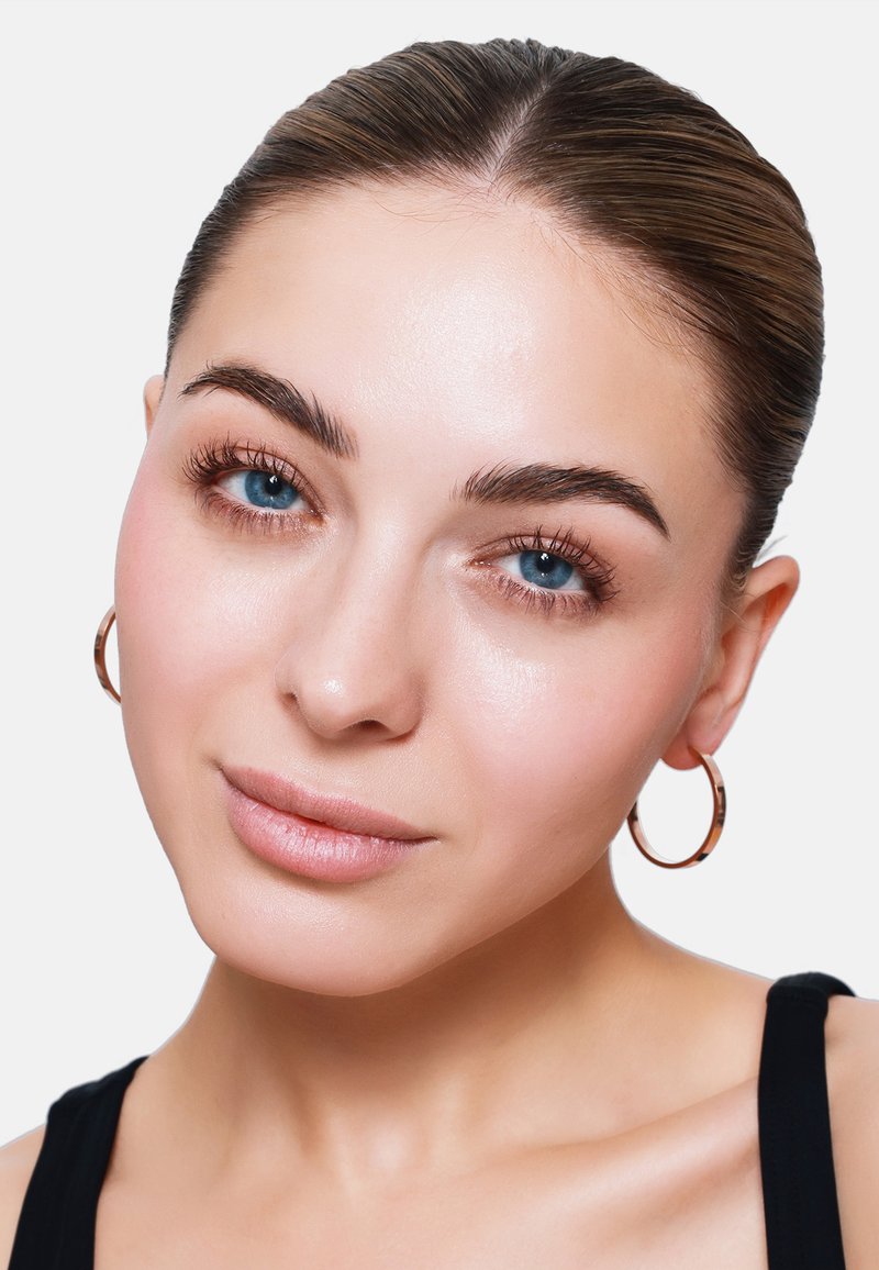 Primo piano di una persona con pelle liscia, occhi blu e trucco leggero, che indossa piccoli orecchini a cerchio d'oro e ha un’acconciatura a chignon elegante.