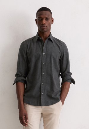 Camicia grigia con bottoni e maniche lunghe, con polsini arrotolabili e una texture sottile. Abbinata a pantaloni di colore chiaro.