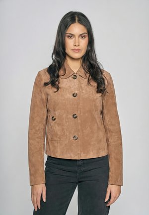 Frau mit langem dunklem Haar, die eine hellbraune, geknöpfte Wildlederjacke und schwarze Hose trägt, vor einem schlichten grauen Hintergrund stehend.