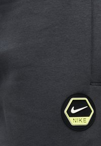 Tessuto grigio con una texture liscia, caratterizzato da una toppa con il logo Nike esagonale nero e giallo sul lato inferiore sinistro.