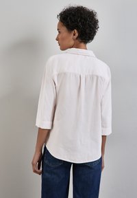 Light pink, long-sleeve shirt with a collar, featuring a back pleat and rolled cuffs, paired with dark blue jeans.