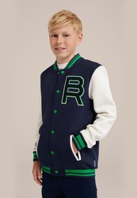 Navyblå och vit varsityjacka med gröna accenter; har en broderad "B" på bröstet, knappar och randiga ribbade muddar och nederkant.