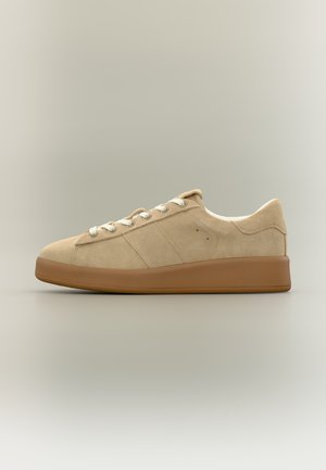 Beżowy zamszowy niski sneaker z białymi sznurowadłami i jasnobrązową gumową podeszwą, przedstawiony z boku na neutralnym tle.