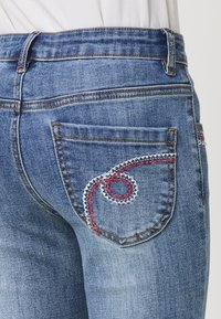 Jeans de mezclilla azul desgastados con bolsillos traseros que presentan un intrincado bordado en blanco y rojo en un patrón en espiral. Cintura estándar y corte clásico.