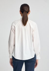 Camicia di cotone bianca con maniche lunghe, caratterizzata da un orlo arrotondato e una vestibilità rilassata. La vista posteriore evidenzia le cuciture delle spalle e una leggera arricciatura nel giromanica.