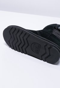 Schwarze Wildlederstiefel mit einer strukturierten Gummisohle, die ein Zickzackmuster und ein geprägtes Logo aufweist. Das Design umfasst Riemen zum Befestigen.